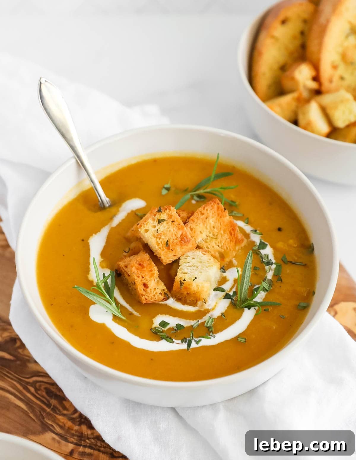 A large white bowl of orange squash soup with croutons, herbs, and coconut milk on top. There is a spoon sitting in the soup.