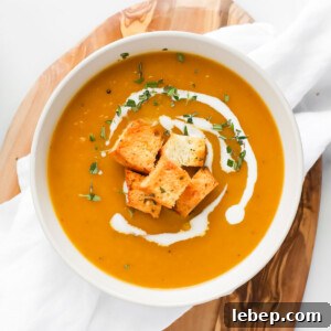 A white bowl filled with orange soup. The soup is garnished with a drizzle of white coconut milk, croutons, and chopped green herbs.