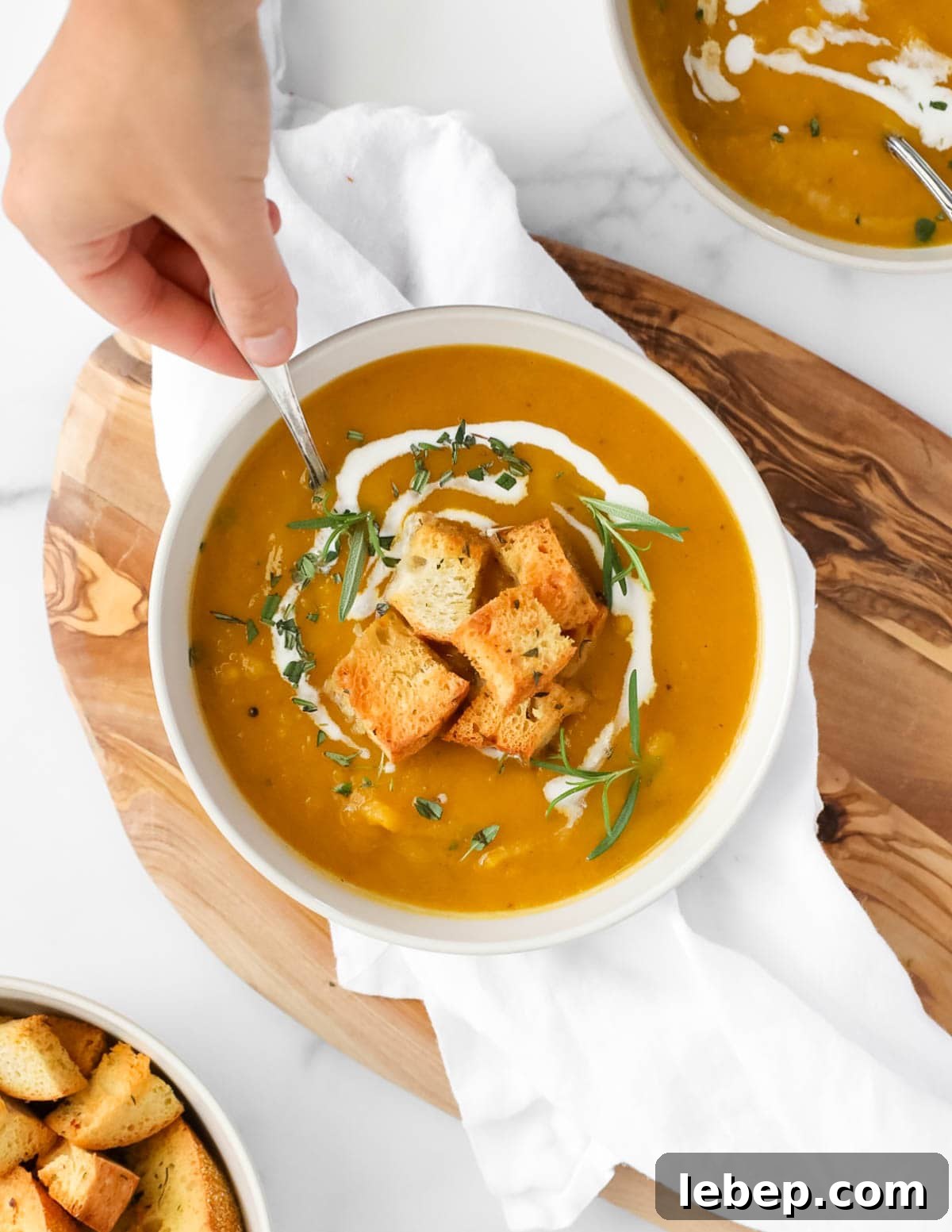 A hand dipping a spoon into a bowl of orange soup. There are herbs, croutons, and coconut milk on top of the soup.