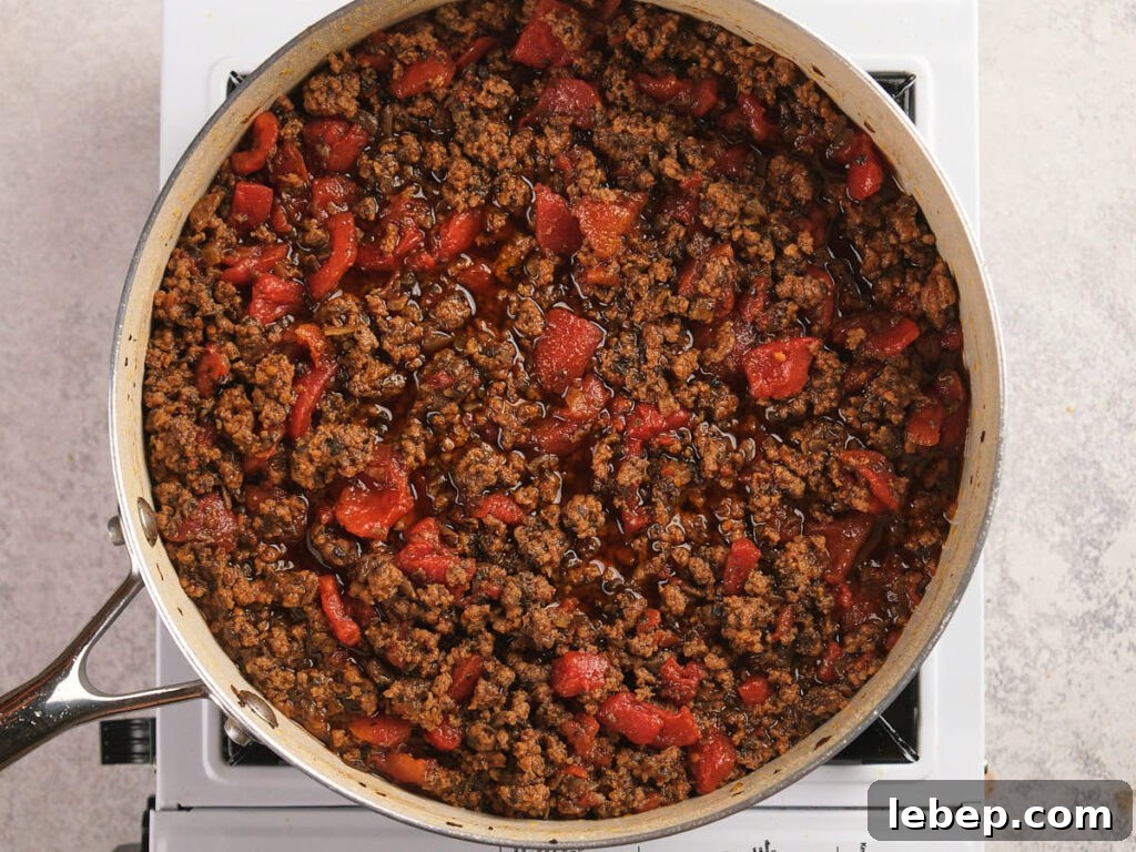A ground beef, mushrooms and diced tomatoes are being simmered in a white skillet.