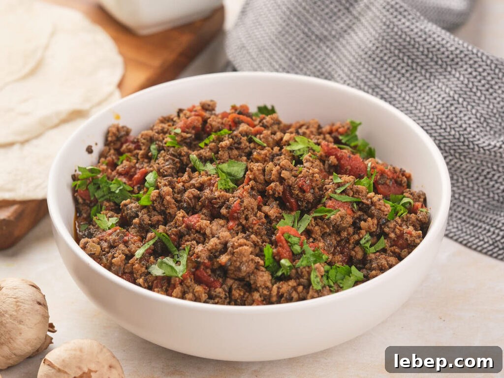 A white bowl of seasoned meat garnished with fresh cilantro alongside with two garlic cloves, tortillas and a table cloth.
