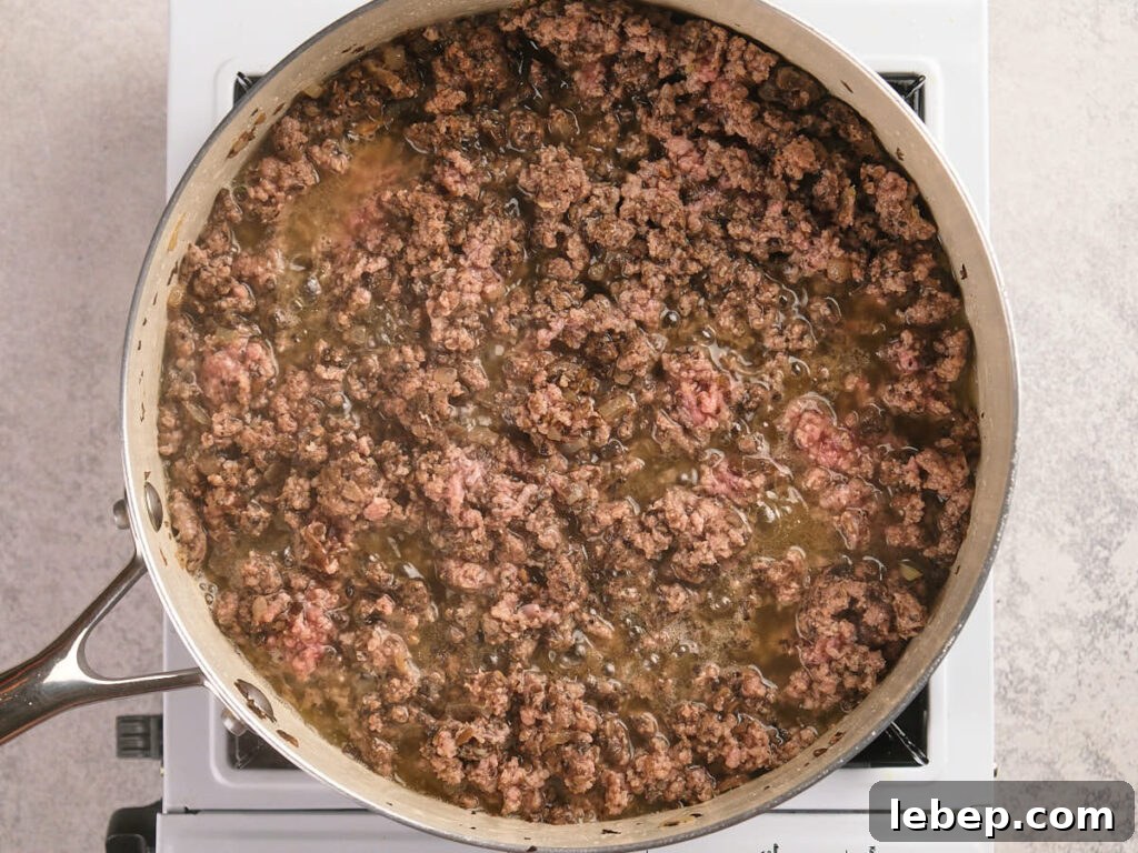 A ground beef is simmering in it's juices on a stovetop, appearing partially cooked in a skillet.