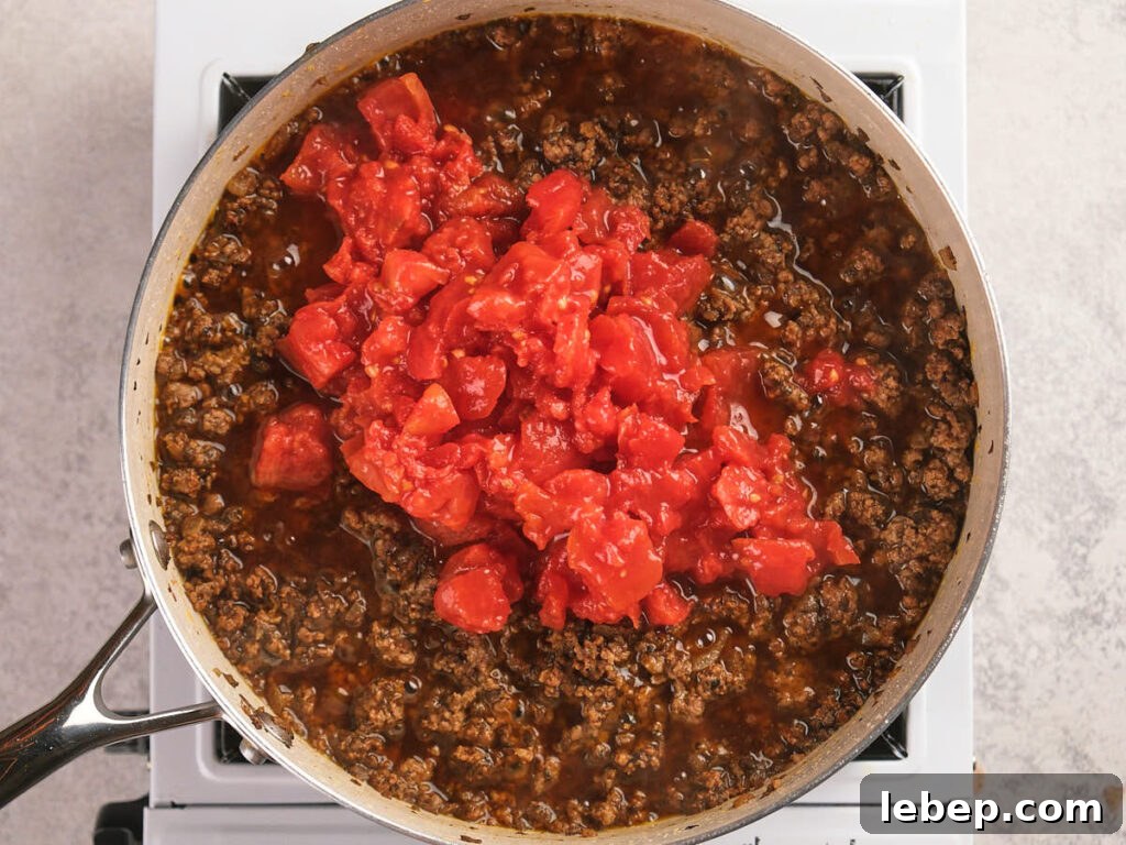 A skillet of cooked ground beef is being simmered with a diced tomatoes added on top.