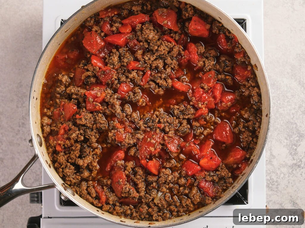A seasoned ground beef and diced tomatoes are combined and being cooked in a white skillet.