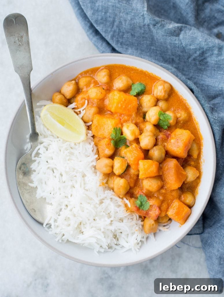 Sweet potato chickpea curry served in a white bowl with a spoon and has fresh cilantro and lemon slice as garnish.