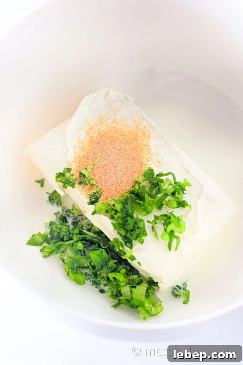 Cheesy Chicken Crescent Bake with Puff Pastry Squares 6 A block of cream cheese topped with green herbs and brown seasoning nestles in a white bowl, serving as an ideal starting point for a delightful recipe or paired perfectly with crescent squares.