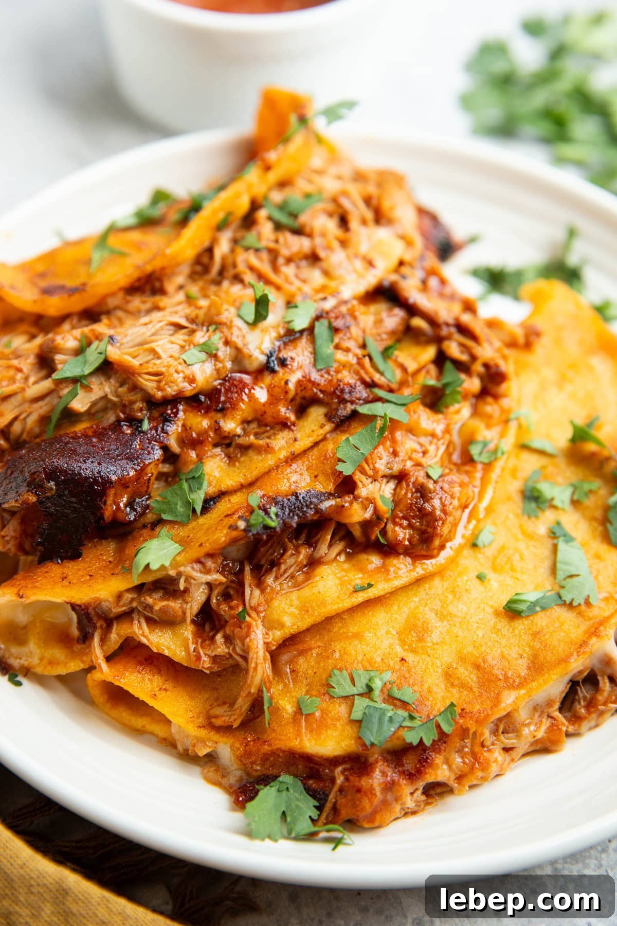 Four crispy corn tortilla tacos on a plate with melted cheese and shredded chicken. Everything sprinkled with fresh cilantro and a bowl of sauce in the background.