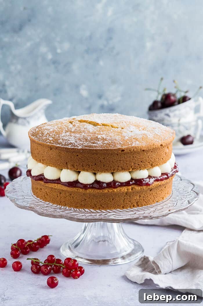 Vegan victoria sponge cake on a glass cake stand with fruit and crockery in the background.